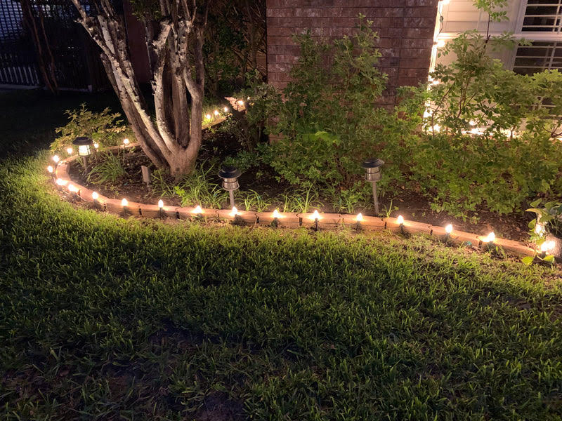 Garden-edge stake lighting Christmas bulbs on stakes follow a curved garden bed beside a home.